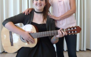 Zwei Personen, eine junge Frau mit Gitarre und eine ältere Frau, sitzen oder stehen zusammen vor einem cremefarbenen Vorhang.