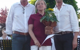 Zwei Männer und eine Frau stehen vor einem grünen Hintergrund mit Kindern im Hintergrund. Die Frau hält ein Blatt Papier.