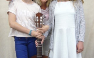 Eine Mutter und ihre Tochter halten zusammen ein Gitarreninstrument. Die beiden tragen weiße Kleidung.