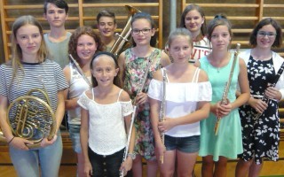 Mehrere junge Menschen halten Blasinstrumente wie eine Tuba, Flöte und Klarinette in der Hand. Sie tragen Shorts und leichte Oberteile.