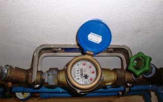 Kalter Wassermesser an einer Wand. Metallisches Gehäuse mit Drehknöpfen und einem kleinen blau-weiß Schild.