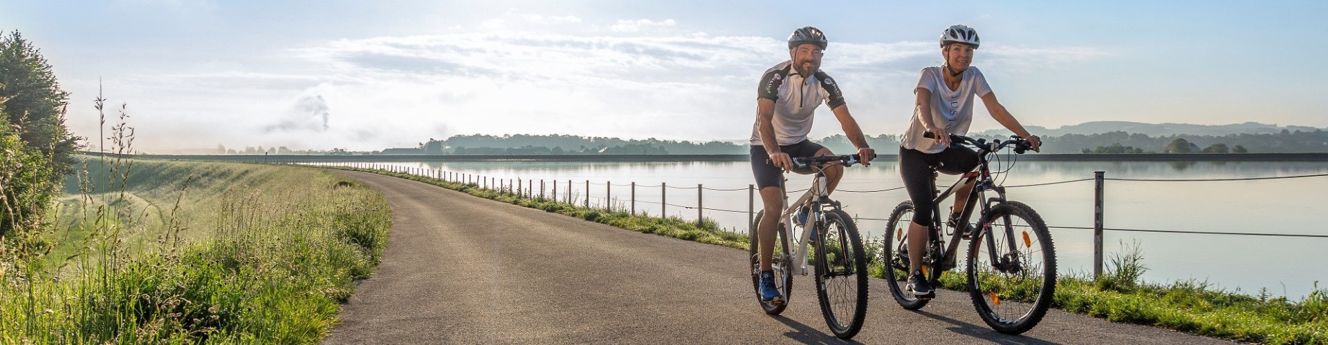 Zwei Radfahrer fahren auf einer asphaltierten Straße entlang eines Sees. Im Hintergrund grasige Hügel und ein Zaun.