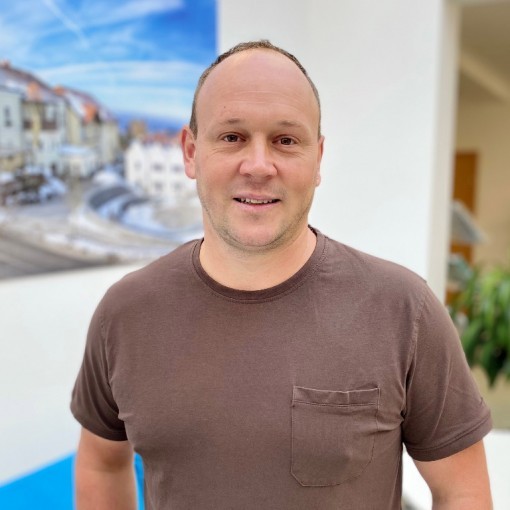 Mann in braunem T-Shirt vor einer blauen Wand mit Stadtbild.