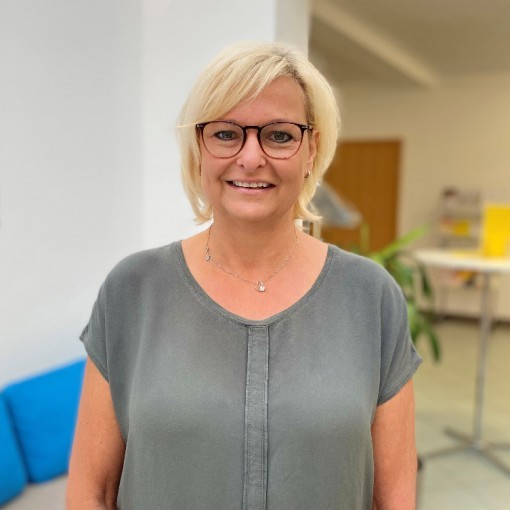 Frau mit braunen Haaren und Brille, die ein schlichtes grünes Kleid trägt. Im Hintergrund ein Schreibtisch und ein blauer Stuhl.