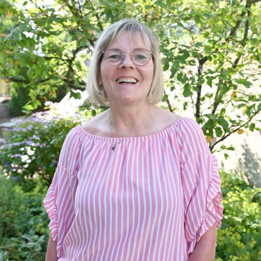Frau in weiß-weiß gestreiftem Oberteil und Jeans, Blick in die Kamera, im Hintergrund Grünpflanzen.