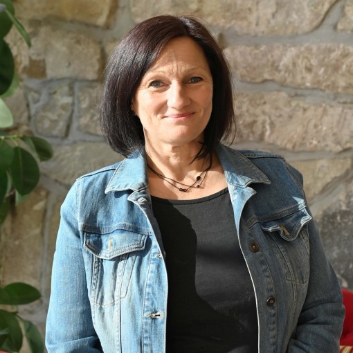 Frau mit dunkler, schulterlanger Mähne trägt blaue Jeansjacke und schwarzen Top. Im Hintergrund roter Sessel und Grünpflanze.