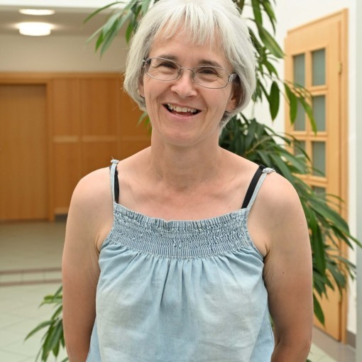 Dame mit blauer Brille und hellblauem Kleid, die freundlich in die Kamera lächelt. Im Hintergrund ein grünes Blattpflanze.