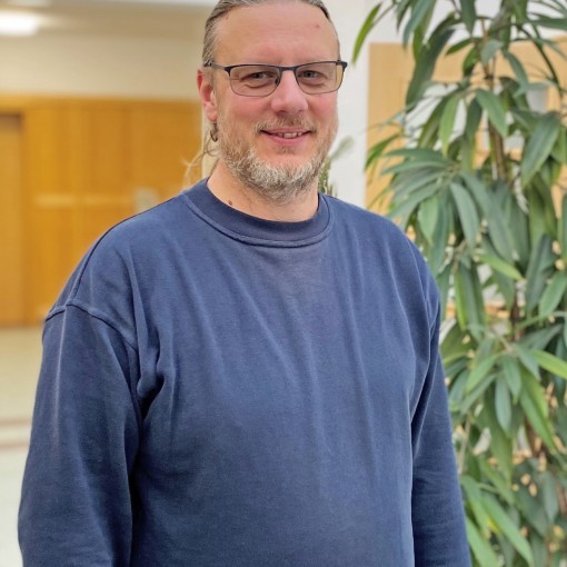 Mann mit braunen Haaren und Brille, gekleidet in einem dunkelblauen Pullover, im Hintergrund eine grüne Zimmerpflanze.