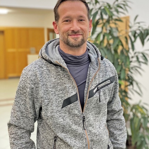 Mann in grauem Strickjacke-Pullover mit Zipfel, gelbliches Hemd, Hände in den Hosentaschen.