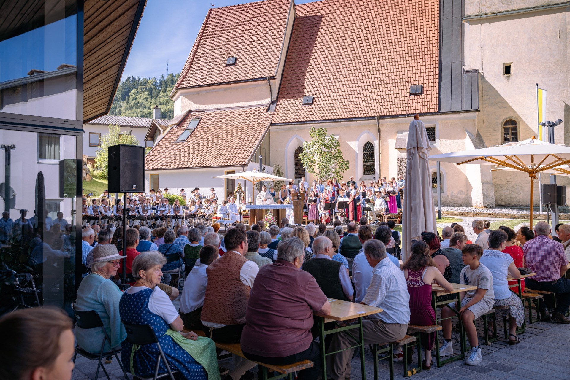 Eine Menschenmenge sitzt auf Bänken vor einem Gebäude mit roten Dächern. Es ist ein Veranstaltungstag mit einer musikalischen Darbietung.