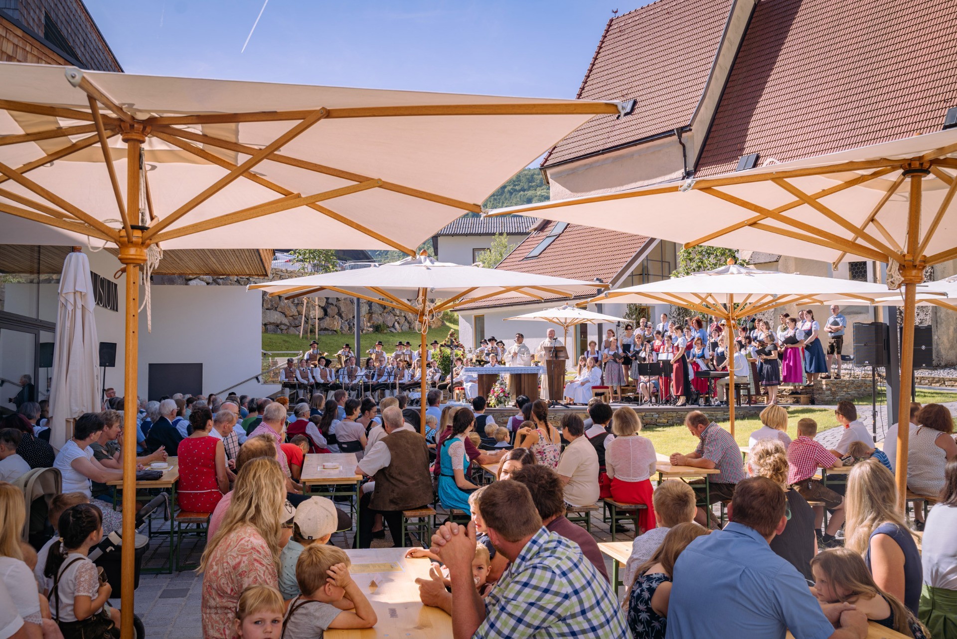 Ein Fest wird gefeiert: Viele Menschen sitzen an Tischen in einem Garten, ein Musikorchester spielt auf einer Bühne.