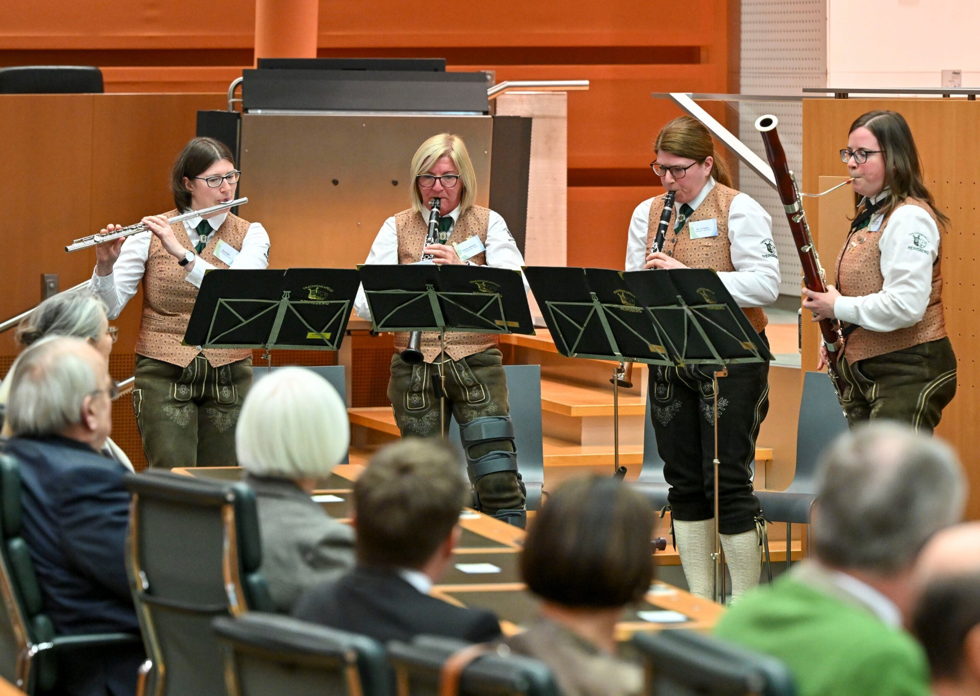Musiker in Trachten spielen auf einer Bühne vor Publikum. Im Vordergrund: Flöten- und Bassflötenspieler.