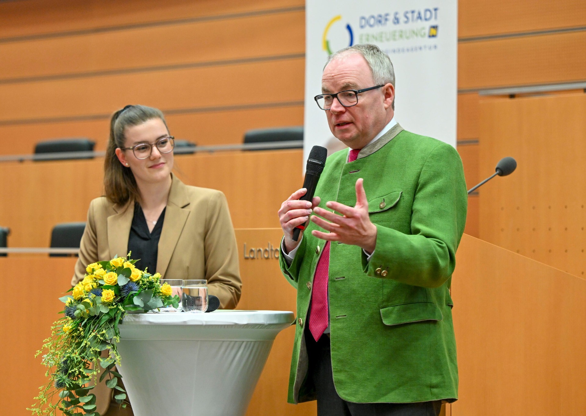 Ein Mann in einem grünen Anzug hält vor einem Sprechmikrofon eine Rede. Im Hintergrund steht eine Frau mit Brille. Auf einem Tisch steht eine Vase mit Blumen.