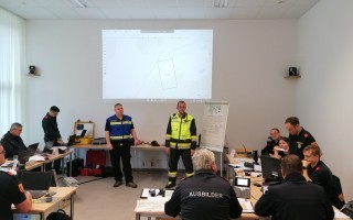 Ein Raum mit mehreren Personen, die an einem Unterrichts- oder Schulungsablauf teilnehmen. Einige Personen tragen Blaulichtuniformen. Ein großer Bildschirm zeigt eine Grafik.