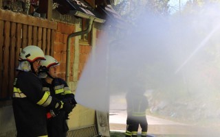 Feuerwehrleute sprühen Wasser bei einem Einsatz. Rauchwolke im Vordergrund, Gebäude im Hintergrund.
