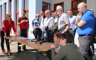 Gruppe von Personen, vermutlich Feuerwehrleute, um einen Tisch in der Nähe eines Gebäudes. Ein Teil der Gruppe blickt auf ein Dokument.