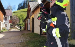 Feuerwehrmann mit Blaulichtuniform sprüht mit einem Wasserschlauch. Im Hintergrund ein Dorfweg und ein Gebäude.