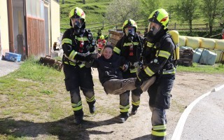 Mehrere Feuerwehrleute tragen einen Verletzten. Sie tragen helle Schutzkleidung mit gelben Akzenten. Im Hintergrund sind Gebäude und Ballaststoffe zu sehen.