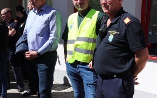 Drei Männer in Arbeitskleidung stehen vor einem Gebäude. Zwei tragen Gelbwesten und schwarze Jacken, der dritte einen dunkelblauen Anzug.