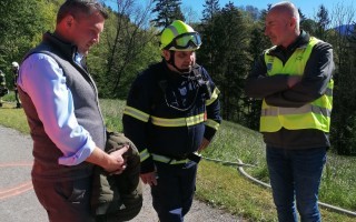 Drei Männer unterhalten sich auf einem Weg, gespickt mit Bäumen. Ein Feuerwehrmann in Warnweste und ein Mann in Mantel schauen auf ein Objekt.