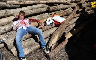 Person liegt blutverschmiert auf einem Haufen Holzstapel. Eine Mütze und ein Plastikbecher sind im Hintergrund zu sehen.