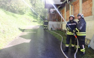 Zwei Feuerwehrleute mit Schutzkleidung spritzen mit einer Schlauchleitung eine Kiesstraße. Im Hintergrund ein Bungalow.