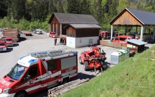 Rotes Feuerwehrauto und ein Radlbagger stehen vor einem einfachen Holzgebäude auf einem Parkplatz.