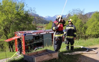 Zwei Feuerwehrleute stehen neben einem roten Feuerwehrauto auf einer grünen Wiese. Ein Wasserrohr fließt aus dem Fahrzeug. Im Hintergrund sind Berge zu sehen.