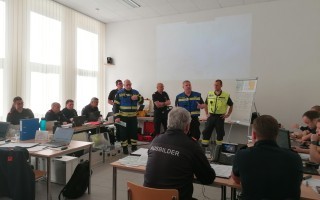 Ein Raum mit mehreren Personen, die an einem Sitzungs- oder Schulungsgespräch teilnehmen. Einige tragen gelbe Westen. Es sind Schreibtische und Computer zu sehen.