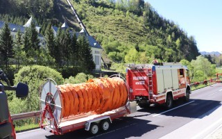 Rotes Feuerwehrauto zieht einen mit orangefarbener Plane beladenen Anhänger auf einer Straße. Im Hintergrund ein Gebäude.