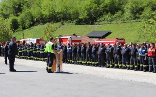 Feuerwehrpersonal steht in einer Reihe und hört einer Person am Rednerpult zu. Im Hintergrund ein Feuerwehrauto.