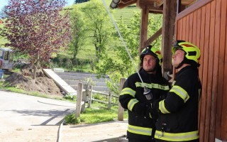 Zwei Feuerwehrleute in gelben Anzügen stehen an einer Wasserleitung neben einem Holzgebäude. Im Hintergrund ein bewachsener Hügel.