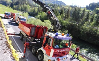 Rotes Feuerwehrfahrzeug mit Krannensystem auf abschottender Straße. Brandbekämpfer in Warnweste steht daneben.