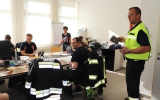 Ein Raum mit mehreren Feuerwehrleuten um einen Tisch. Einer hält ein Tablet hoch, die anderen schauen konzentriert darauf.