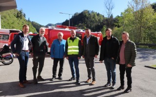 Verschiedene Männer stehen an einer Straße, im Hintergrund ein roter Notfallwagen und ein grüner Wald.