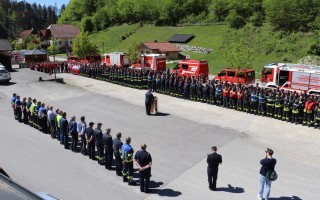 Eine Reihe von Feuerwehrleute steht auf einem Parkplatz vor einem roten Feuerwehrfahrzeug. Im Hintergrund ein Gebäude und weitere Fahrzeuge.