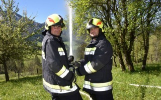 Zwei Feuerwehrleute in schwarze Uniformen mit gelben Sicherheitswesten halten Regenschläuche. Im Hintergrund verschwommen eine grüne Wiese mit Bäumen.