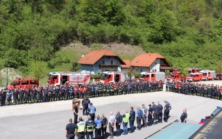 Feuerwehrpersonal steht in einer Reihe vor einem Gebäude mit roten Dächern und Feuerwehrautos.