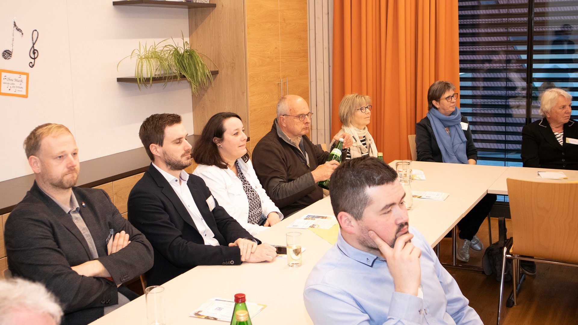 Mehrere Personen sitzen an einem Tisch, vermutlich bei einer Besprechung oder Veranstaltung. Ein Mann mit blauer Jacke und eine Frau mit Brille sind deutlich sichtbar.