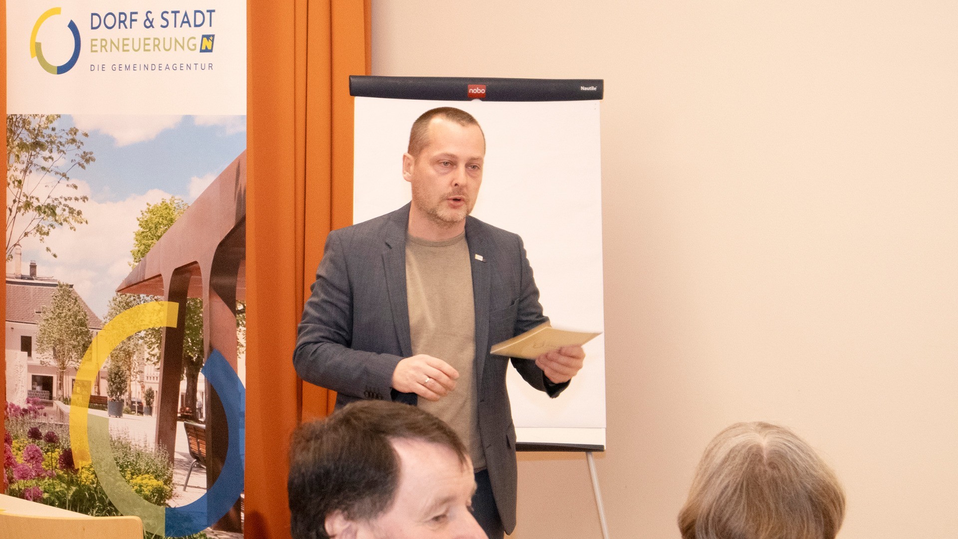 Ein Mann in Anzug steht vor einem schwarzen Display-Rahmen mit einem Landschaftsbild eines Daches und einer Spielgerüst.