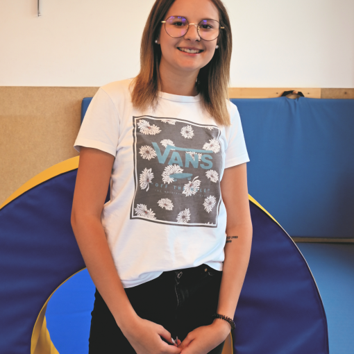 Frau mit Brille, die in die Kamera schaut, trägt ein weißes Shirt mit Vans-Logo und dunkelblaue Jeans. Im Hintergrund ein blau-gelbes Segelcloth.