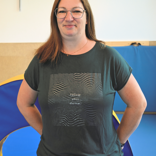 Frau mit braunem Haar, die eine dunkelgrüne T-Shirt mit Wellenmuster trägt, im Hintergrund blaue Hängesessel.