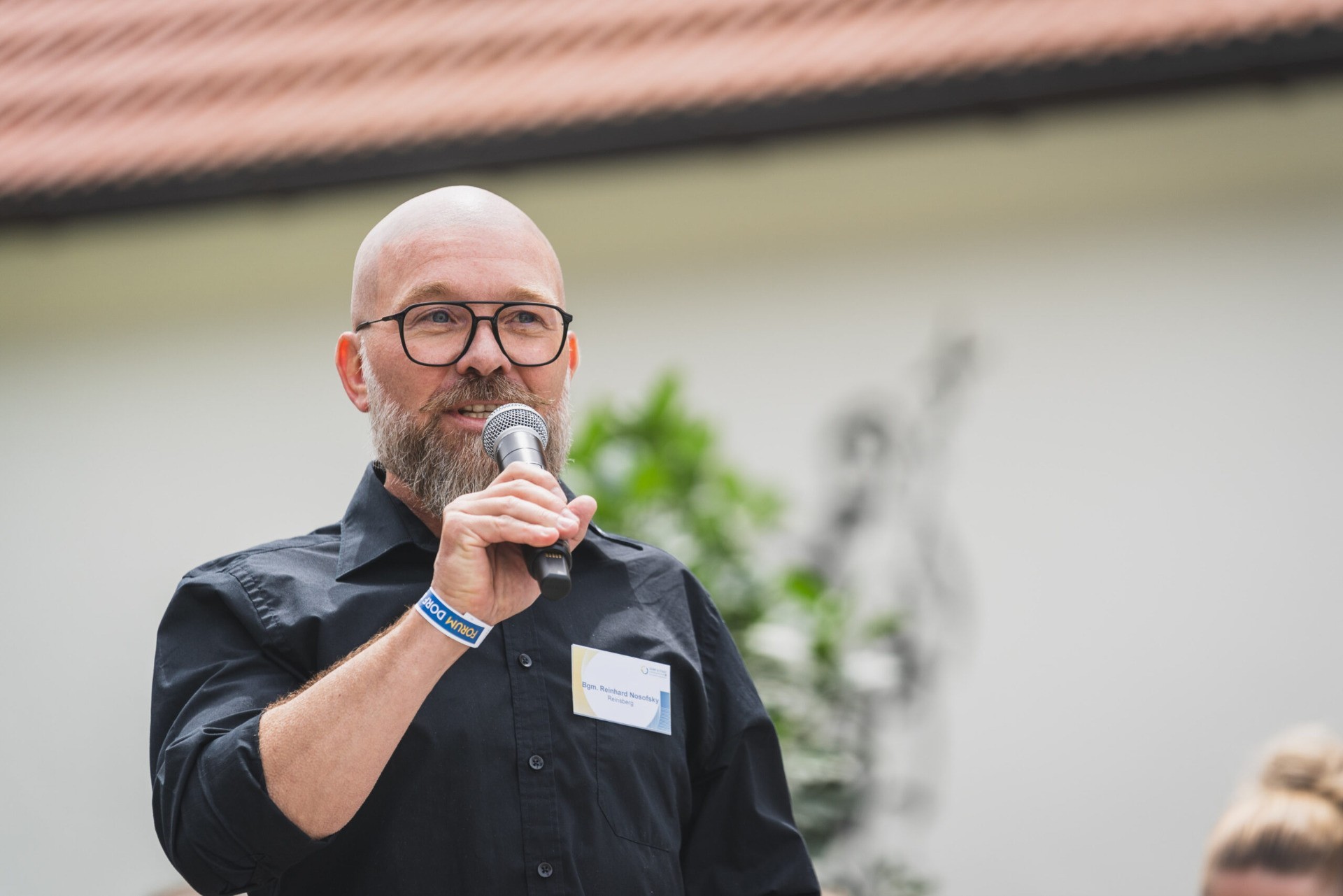 Mann mit Brille spricht in ein Mikrofon, im Hintergrund Grünpflanzen.