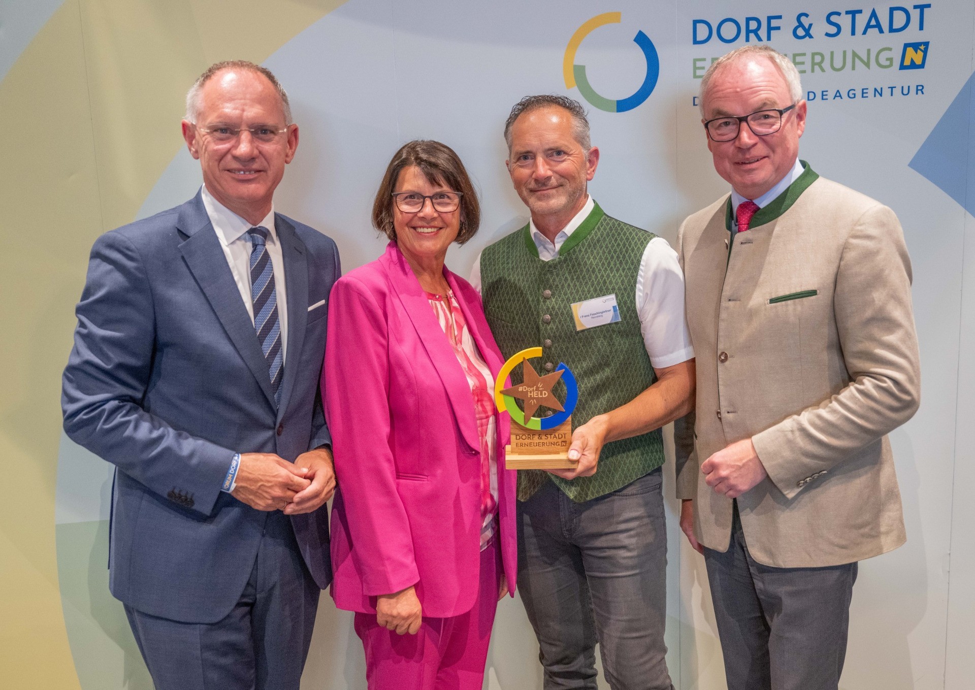 Drei Personen stehen zusammen. Ein Mann in blau, eine Frau in pink und ein Mann in Beige halten einen goldenen Preis.