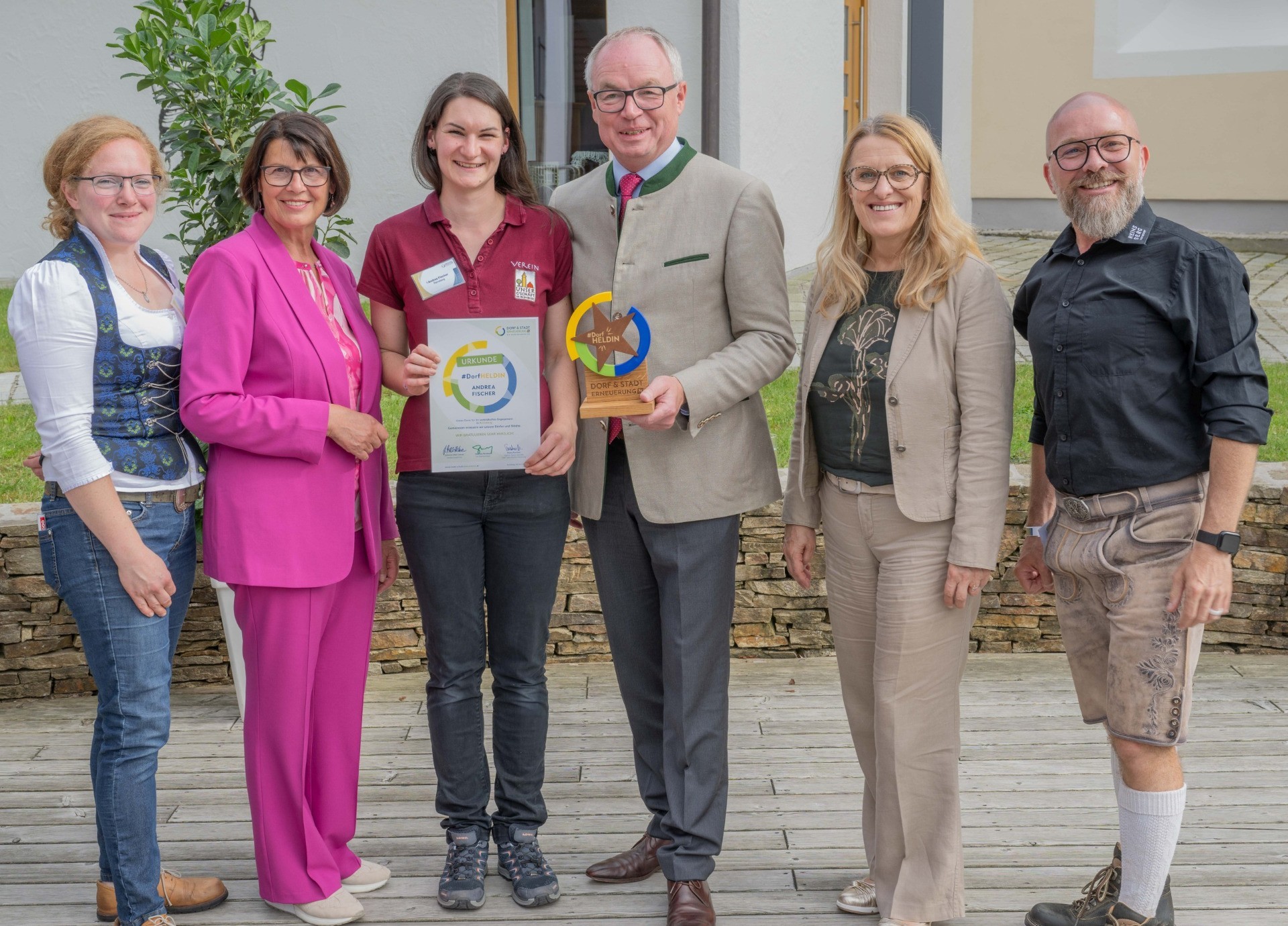 Six people in traditional Bavarian clothing stand outdoors. A woman wears a pink shirt, while others are in brown and grey patterns.