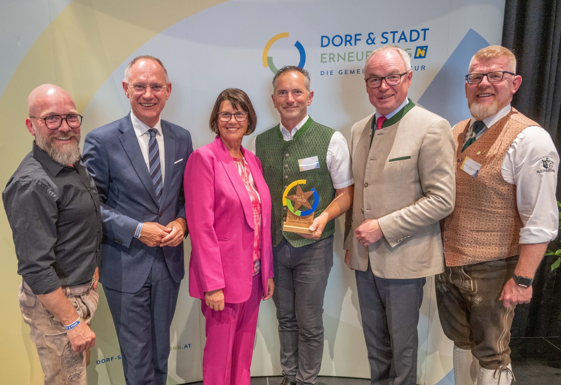 Drei Personen in Trachten: ein Mann und eine Frau in braunen und pinken Trachtenwesten, sowie ein Mann in einer grünen Jacke.