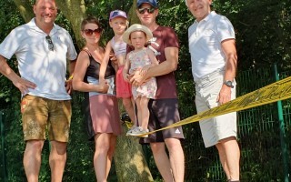 Eine Familie von sechs Personen steht auf einer Wiese. Ein Mann mit Brille, ein Mann mit Sonnenbrille, ein Mann in Shorts und eine Frau mit Kind stehen eng beieinander.