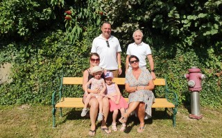 Familie lächelt auf einer Parkbank. Ein Mann, eine Frau und zwei kleine Kinder sitzen zusammen. Im Hintergrund ein Feuerlöscher.