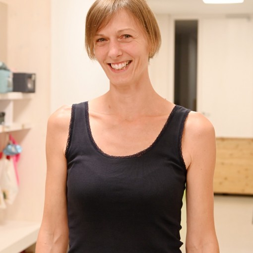 Frau mit braunen Haaren trägt ein schwarzes Tanktop. Im Hintergrund sind Möbel und ein Regal mit Gegenständen zu sehen.