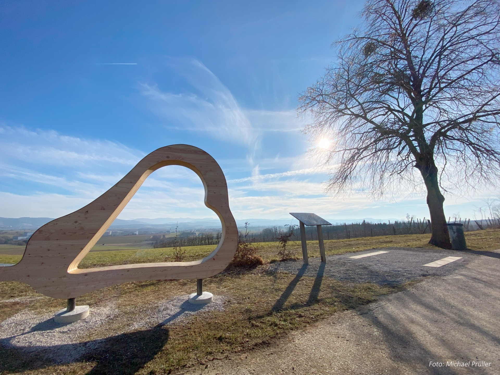 Hügelpanoramabild mit Holzherz-Skulptur und Baum. Im Hintergrund verschwommenes Tal und Wolken am blauen Himmel.